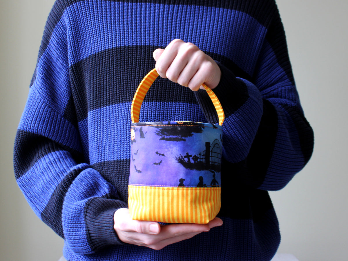 Small Halloween trick or treat basket held by hand, featuring a colorful Halloween print, purple base and contrasting handle.